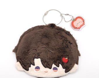May include: A plush brown character-shaped coin purse with a silver keyring and a small apple charm. The coin purse has a smiling face with purple eyes and a small red apple detail. The apple charm has a smiling face.