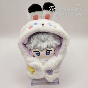 May include: A plush doll wearing a white hooded bunny outfit with black and pink accents. The doll has gray hair and blue eyes. The hood is buttoned with a purple button. The text "The Deepspace Dream Shop" is in the upper right corner.