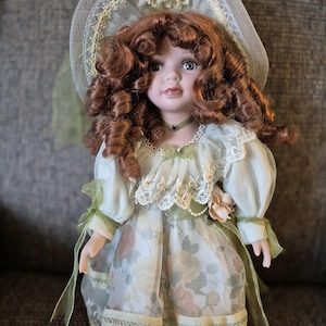 May include: A porcelain doll with auburn curly hair, wearing a light green dress with a floral pattern and lace trim. The doll has a matching hat and a green ribbon around its neck. The doll is a collectible item.