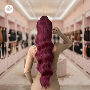 May include: A long, wavy, burgundy-colored ponytail hair extension. The hair is styled in a high ponytail, with long, flowing waves. The background is a beauty salon with various hair extensions on display.