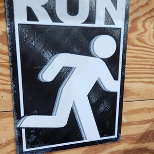 May include: A rectangular sign with the word "RUN" in white block letters above a white silhouette of a person running. The background is black, and the sign has a white border. The sign is mounted on a wooden surface.