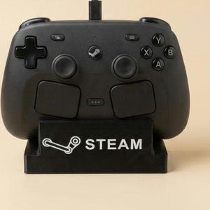 May include: A black video game controller with two analog sticks, a directional pad, and four action buttons labeled A, B, X, and Y. The controller rests on a black stand featuring the white Steam logo and the text STEAM printed on the front.