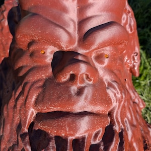 Op de afbeelding: Een close-up van een roodbruine sculptuur van het gezicht van een Bigfoot-achtig wezen. De sculptuur heeft een textuur, prominente wenkbrauwen, een grote neus en kleine, oranje ogen. De achtergrond is wazig groen gras.