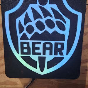 以下が含まれることがあります： 黒い長方形の看板。盾のデザイン、クマの爪の跡、白い文字で「BEAR」と書かれています。盾とクマの爪は青と緑のグラデーションです。黒いコードが下から伸びています。