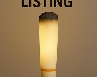 May include: A quirky lamp designed to resemble a giant cigarette. It features a dark brown top, an off-white body with an orange band, and a warm, internal glow. The words "RESERVED LISTING" are displayed above.