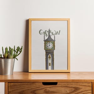 May include: A framed print featuring a detailed illustration of the Gastown steam clock. The clock tower is gray and green with a clock face and the word "Gastown" above it. The print is displayed on a wooden surface.