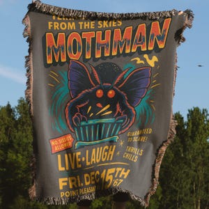 May include: Dark gray woven throw blanket featuring a Mothman design with the text "FROM THE SKIES MOTHMAN". The design includes a cartoon Mothman, the text "LIVE LAUGH", and "FRI. DEC 15TH '67". The blanket has fringed edges.