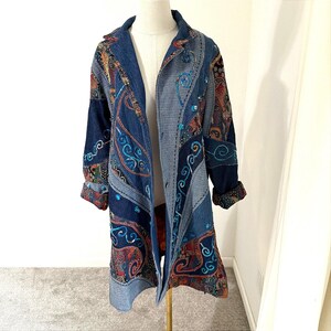 Pode incluir: Um casaco de ganga de manga comprida com um design de patchwork, apresentando vários tons de azul, painéis de tecido escuro com padrões paisley intrincados e decorações de lantejoulas turquesa ao longo das costuras e painéis frontais.