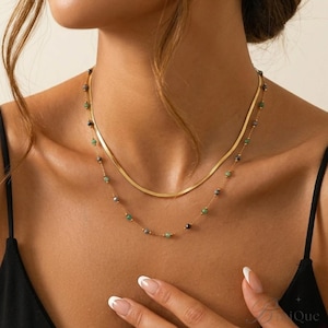 May include: Two gold necklaces. The top necklace is a simple, flat chain. The second necklace has small, colorful beads in shades of green, black, and gray. The necklaces are worn on a person's neck, with a black top.