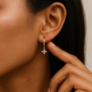 Peut inclure: Boucle d'oreille dorée avec un pendentif en forme de fleur à quatre pétales, chaque pétale serti d'une petite pierre précieuse transparente. La boucle d'oreille est portée à l'oreille, mettant en valeur les détails délicats et l'éclat du bijou.