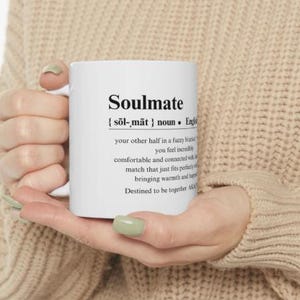 May include: White ceramic mug with a black and white definition of the word "Soulmate". The definition reads: "{sol-mat} noun. English. your other half in a fuzzy blanket. You feel incredibly comfortable and connected with a match that just fits perfectly, bringing warmth and happiness. Destined to be together. Always."