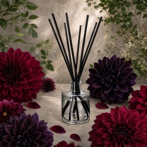 May include: A clear glass reed diffuser bottle filled with liquid and containing several thin black reeds arranged in a fan shape. The bottle is surrounded by multiple large, deep red and dark purple dahlia flowers with layered petals.