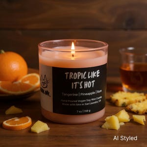 May include: A lit, pink-colored candle in a clear glass jar with the text "Tropic Like It's Hot". The candle is scented with tangerine, pineapple, and rum. Orange slices and pineapple chunks are arranged around the candle.