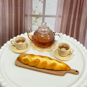 May include: A miniature tea set with a glass teapot, two teacups with saucers, and a loaf of bread on a wooden cutting board. The tea is amber-colored, and the cups have lemon slices. The set is on a white, scalloped tray.