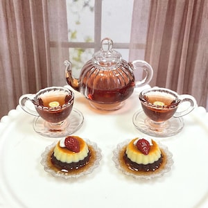May include: A miniature tea set with a glass teapot, two teacups, saucers, and two desserts. The teapot and teacups are filled with tea. The desserts have a white topping and a strawberry on top. The set is on a white tray.