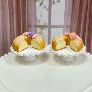 May include: Two miniature bundt cakes, each cut to reveal the interior, sit on white scalloped cake stands. The cakes are decorated with pink and purple flower accents. The cakes are on a white serving platter.