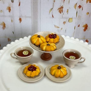 May include: A miniature tea set with pumpkin-shaped pastries and two cups of tea, each with a lemon slice. The pastries are on a tiered serving dish and small plates. The tea cups are light beige.