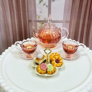 May include: A miniature tea set with a pink glass teapot, two teacups, and a tiered serving tray with pastries. The tea cups and teapot are filled with a reddish-brown liquid. The serving tray holds miniature cakes and pastries.