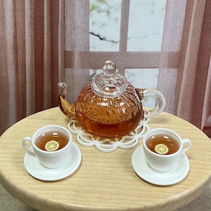 May include: A miniature tea set featuring a clear glass teapot filled with tea, two white teacups with lemon slices, and white saucers. The teapot sits on a white decorative coaster. The tea set is on a small wooden table.