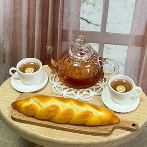 May include: A miniature amber-colored glass teapot with a lid, two white ceramic teacups on matching saucers filled with tea and lemon slices, and a braided loaf of bread on a wooden serving board, all arranged on a small round table.