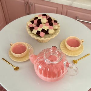 May include: A pink glass teapot with a clear lid, two pink teacups with saucers, and a plate of pink and white checkered cookies. The teacups and teapot are filled with pink liquid.