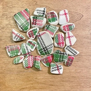 May include: A collection of white, irregularly shaped stones, each adorned with a plaid pattern in red, green, and black. The stones are scattered on a light brown wooden surface, creating a rustic aesthetic.