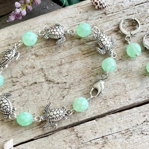 以下が含まれることがあります： シルバーカラーのブレスレットと、お揃いのイヤリングのセット。ブレスレットには小さなカメのチャームと、薄い緑色のファセットビーズが付いています。イヤリングにもお揃いの緑色のビーズが付いています。ジュエリーは、風化した木製の表面に展示されています。