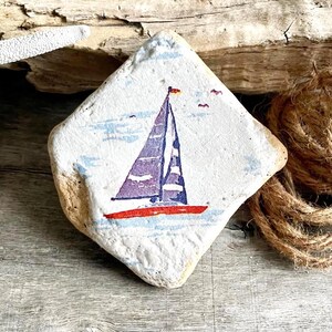 May include: A diamond-shaped, hand-painted stone features a sailboat with a purple sail and red hull. The artwork includes blue water and sky, with small birds. The stone has a natural, textured appearance.