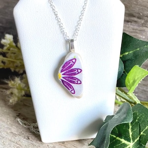 May include: A silver chain necklace with a pendant featuring a white background and a purple flower design. The flower has a yellow center. The necklace is displayed on a white jewelry stand.