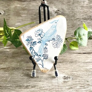 以下が含まれることがあります： 青い鳥と花の模様が描かれた、白い三角形の陶器。鳥は花のある枝にとまっています。陶器は、木製の背景に対して黒い金属製のスタンドに展示されています。