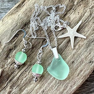 May include: A sea-themed jewelry set featuring a necklace with a light green sea glass pendant, matching earrings with light green beads, and a starfish charm. The set is made of silver-colored metal and rests on a wooden surface.