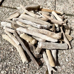 May include: A pile of weathered driftwood pieces in various shapes and sizes. The wood is primarily light brown and gray, with some pieces showing darker, weathered textures. The pieces are arranged on a textured, gray surface.