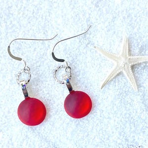 May include: A pair of red, round earrings with silver-colored hooks and accents. The earrings are displayed on a white, textured surface with a white starfish in the background. The earrings have a matte finish.