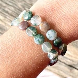 May include: Two beaded bracelets with round, polished stones in shades of green, white, and brown. The bracelets are worn on a wrist, showcasing the natural variations in the stone colors and patterns. The beads are approximately 0.5 inches in diameter.