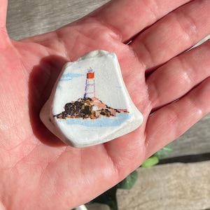 Puede incluir: Un pequeño trozo de cerámica blanco de forma irregular con una escena de faro pintada. El faro tiene una parte superior roja y está situado en una isla rocosa con agua y cielo azules. La obra de arte es detallada y colorida.
