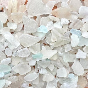 Puede incluir: Una colección de pequeñas piezas de vidrio marino esmerilado y liso en varios tonos de blanco, transparente, rosa pálido y azul aguamarina claro. Los fragmentos tienen bordes redondeados y desgastados con una textura mate y translúcida.