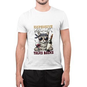May include: White t-shirt featuring a graphic of a cat wearing sunglasses and a bandana, holding a glass of wine and a bottle of wine. The text on the shirt reads "Expensive, difficult, talks backs."