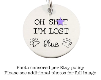 May include: A silver dog tag with the text "OH SHAT I'M LOST" and the word "Blue" printed on it. There are two paw prints on either side of the word "Blue".