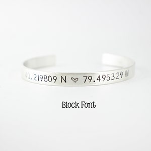 May include: A silver-toned cuff bracelet featuring the engraved coordinates 40.219809 N ♡ 79.495329 W. The text Block Font is printed below the bracelet. The bracelet has an open design with a smooth, metallic finish and clean, straight edges.