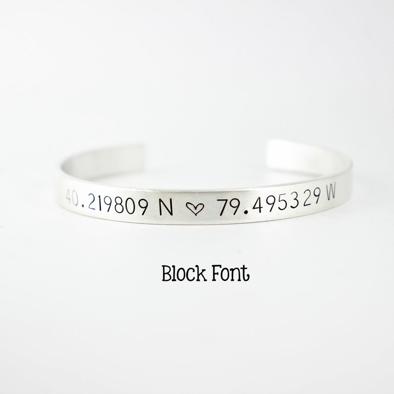 May include: A silver-toned cuff bracelet featuring the engraved coordinates 40.219809 N ♡ 79.495329 W. The text Block Font is printed below the bracelet. The bracelet has an open design with a smooth, metallic finish and clean, straight edges.