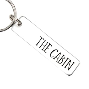 Könnte beinhalten: Ein silberner Schlüsselanhänger mit einem rechteckigen Anhänger, auf dem in schwarzen Buchstaben "THE CABIN" steht.