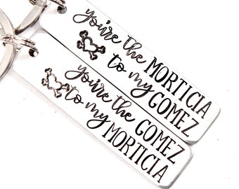 May include: Two rectangular silver-toned metal keychains with split rings. One keychain is engraved with "You're the MORTICIA to my GOMEZ" and a heart-shaped skull icon. The other is engraved with "You're the GOMEZ to my MORTICIA" and a heart-shaped skull icon.