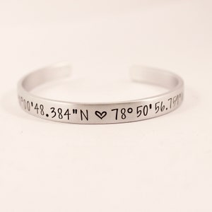 May include: A silver-toned metal cuff bracelet with an open back. The exterior surface is engraved with black text reading 00'48.384" N, a small heart symbol, and 78&deg; 50' 56.759" W. The bracelet has a smooth, matte finish and a simple, curved design.