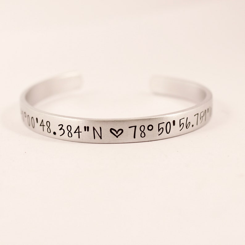 May include: A silver-toned metal cuff bracelet with an open back. The exterior surface is engraved with black text reading 00'48.384" N, a small heart symbol, and 78&deg; 50' 56.759" W. The bracelet has a smooth, matte finish and a simple, curved design.