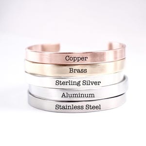 May include: A stack of five metal cuff bracelets with a smooth finish. Each bracelet is engraved with its material name: Copper, Brass, Sterling Silver, Aluminum, and Stainless Steel. The bracelets are arranged in a vertical column on a white surface.
