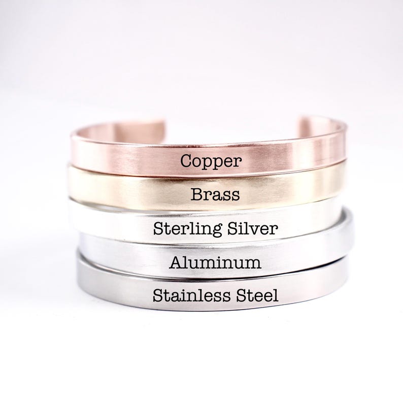 May include: A stack of five metal cuff bracelets with a smooth finish. Each bracelet is engraved with its material name: Copper, Brass, Sterling Silver, Aluminum, and Stainless Steel. The bracelets are arranged in a vertical column on a white surface.