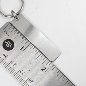 May include: A silver keychain with a blank rectangular tag. The tag is measured against a ruler, showing it is about 2 inches long.