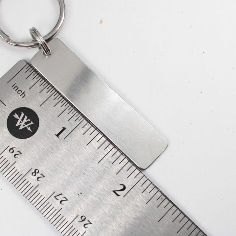 May include: A silver keychain with a blank rectangular tag. The tag is measured against a ruler, showing it is about 2 inches long.