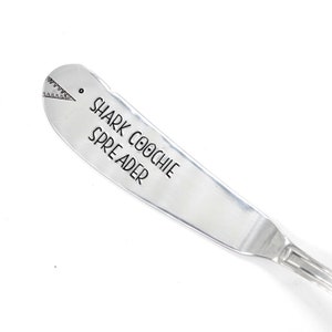 May include: A silver butter knife with a shark fin design and the text "SHARK COOCHI SPREADER" engraved on the handle.