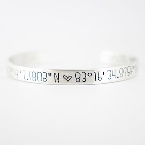 May include: A silver-colored metal cuff bracelet featuring engraved black text that reads 42&deg;14'7.1808" N ♡ 83&deg;16'34.8954" N. The bracelet has a smooth, matte finish and a simple, open-ended circular design that curves gently across the frame.